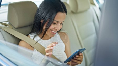 Foto de Joven hermosa mujer hispana pasajero usando teléfono inteligente sentado en el coche en la calle - Imagen libre de derechos
