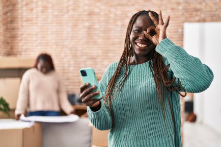 Foto de Mujer africana usando el teléfono inteligente en el nuevo hogar sonriendo feliz haciendo signo bien con la mano en el ojo mirando a través de los dedos - Imagen libre de derechos