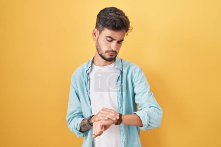 Téléchargez les photos : Jeune homme hispanique avec des tatouages debout sur fond jaune vérifier l'heure sur la montre-bracelet, détendu et confiant - en image libre de droit