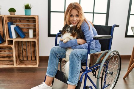 Téléchargez les photos : Jeune femme caucasienne assise sur un fauteuil roulant étreignant chien à la maison - en image libre de droit