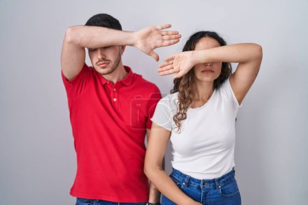 Téléchargez les photos : Jeune couple hispanique debout sur fond isolé couvrant les yeux avec le bras, l'air grave et triste. concept de non-visibilité, de dissimulation et de rejet - en image libre de droit