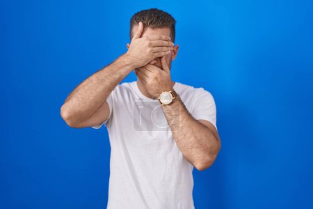 Photo for Young caucasian man standing over blue background covering eyes and mouth with hands, surprised and shocked. hiding emotion - Royalty Free Image
