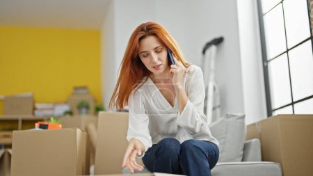 Téléchargez les photos : Jeune rousse femme déballage boîte en carton parler sur smartphone regardant bouleversé à la nouvelle maison - en image libre de droit