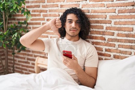 Téléchargez les photos : Homme hispanique avec les cheveux bouclés en utilisant smartphone assis sur le lit avec le visage en colère, signe négatif montrant aversion avec les pouces vers le bas, concept de rejet - en image libre de droit