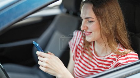 Téléchargez les photos : Jeune femme blonde utilisant smartphone assis sur la voiture à la rue - en image libre de droit