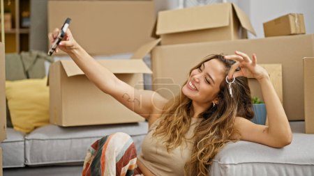 Foto de Joven hermosa mujer hispana sonriendo sosteniendo nuevas llaves de la casa tomando selfie en nuevo hogar - Imagen libre de derechos