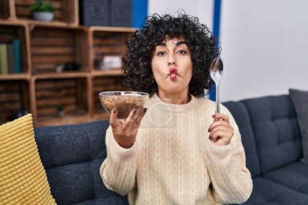 Photo for Young brunette woman with curly hair eating healthy whole grain cereals with spoon making fish face with mouth and squinting eyes, crazy and comical. - Royalty Free Image