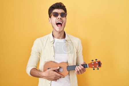 Foto de Joven hombre hispano jugando ukelele sobre fondo amarillo enojado y loco gritando frustrado y furioso, gritando con ira mirando hacia arriba. - Imagen libre de derechos