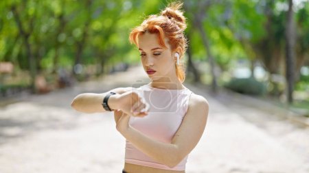 Foto de Mujer pelirroja joven con ropa deportiva y auriculares mirando el reloj en el parque - Imagen libre de derechos