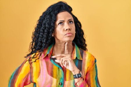 Téléchargez les photos : Moyen Age femme hispanique debout sur fond jaune pensée concentrée sur le doute avec le doigt sur le menton et la recherche jusqu'à se demander - en image libre de droit