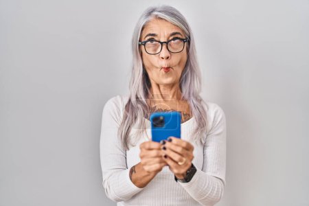Foto de Mujer de mediana edad con cabello gris utilizando el mensaje de escritura de teléfonos inteligentes haciendo cara de pez con la boca y los ojos entrecerrados, loco y cómico. - Imagen libre de derechos