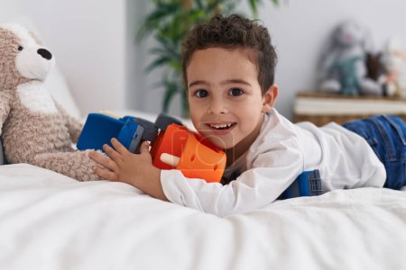 Téléchargez les photos : Adorable garçon hispanique jouant avec jouet de voiture couché sur le lit dans la chambre - en image libre de droit