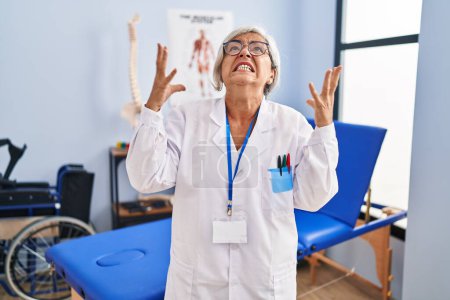 Foto de Mujer de mediana edad con el pelo gris trabajando en la clínica de recuperación del dolor loca y loca gritando y gritando con expresión agresiva y los brazos levantados. concepto de frustración. - Imagen libre de derechos