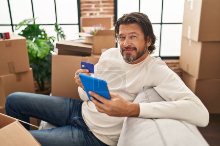 Foto de Hombre de mediana edad utilizando touchpad y tarjeta de crédito sentado en el sofá en el nuevo hogar - Imagen libre de derechos