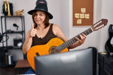 Téléchargez les photos : Femme hispanique du Moyen Âge jouant de la guitare classique au studio de musique souriant heureux et positif, pouce levé faire excellent et signe d'approbation - en image libre de droit