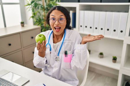Téléchargez les photos : Jeune médecin hispanique femme tenant pomme verte célébrant la réalisation avec sourire heureux et expression gagnante avec la main levée - en image libre de droit