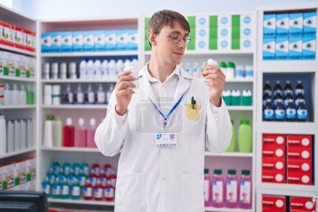 Téléchargez les photos : Young caucasian man pharmacist smiling confident holding pills bottles at pharmacy - en image libre de droit