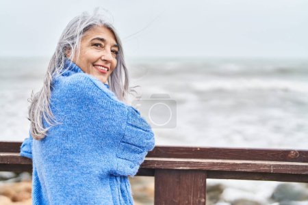 Foto de Mujer de pelo gris de mediana edad sonriendo confiada de pie a la orilla del mar - Imagen libre de derechos