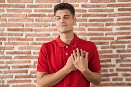 Téléchargez les photos : Jeune homme hispanique debout sur un mur de briques souriant avec les mains sur la poitrine avec les yeux fermés et geste reconnaissant sur le visage. concept de santé. - en image libre de droit