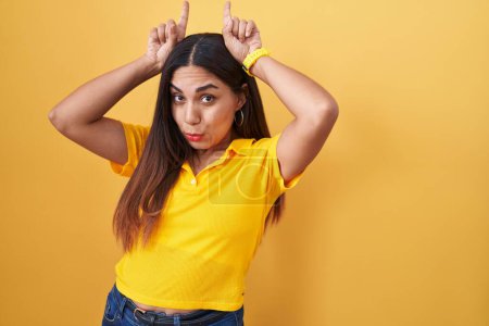 Téléchargez les photos : Jeune femme arabe debout sur fond jaune faisant un geste drôle avec le doigt sur la tête comme des cornes de taureau - en image libre de droit