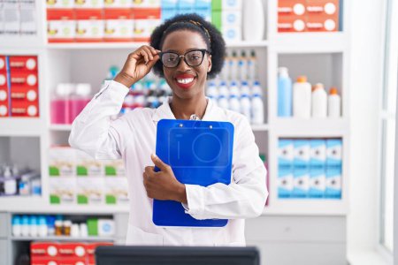 Téléchargez les photos : Afro-américaine femme pharmacien sourire confiant tenant presse-papiers à la pharmacie - en image libre de droit