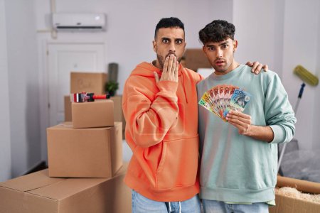 Photo for Young hispanic gay couple moving to a new home holding banknotes covering mouth with hand, shocked and afraid for mistake. surprised expression - Royalty Free Image