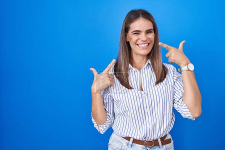 Foto de Mujer joven hispana de pie sobre fondo azul sonriendo alegre mostrando y señalando con los dedos los dientes y la boca. concepto de salud dental. - Imagen libre de derechos