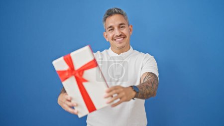 Photo for Young hispanic man smiling confident holding gift over isolated blue background - Royalty Free Image
