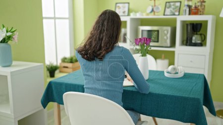 Foto de Joven mujer hispana hermosa sentada en la mesa al revés ver la escritura en el cuaderno en casa - Imagen libre de derechos