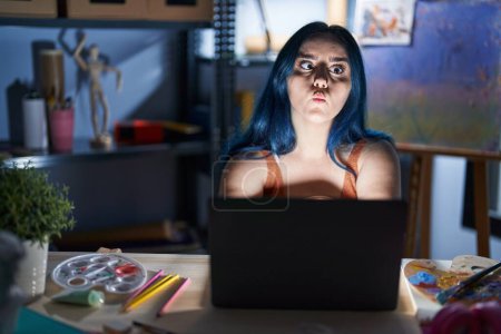 Photo for Young modern girl with blue hair sitting at art studio with laptop at night making fish face with lips, crazy and comical gesture. funny expression. - Royalty Free Image