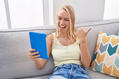 Photo for Young caucasian woman using touchpad sitting on the sofa pointing thumb up to the side smiling happy with open mouth - Royalty Free Image