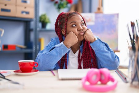 Téléchargez les photos : Artiste afro-américaine assise sur la table avec une expression malheureuse au studio d'art - en image libre de droit