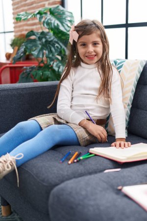 Foto de Adorable chica hispana dibujando en un cuaderno sentado en un sofá en casa - Imagen libre de derechos
