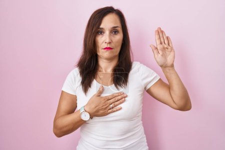Photo for Middle age brunette woman standing over pink background swearing with hand on chest and open palm, making a loyalty promise oath - Royalty Free Image