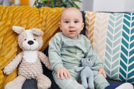 Téléchargez les photos : Adorable bébé caucasien assis sur un canapé avec des poupées à la maison - en image libre de droit