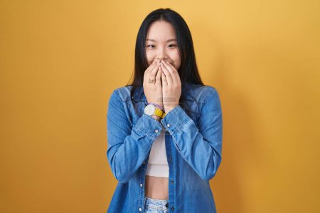 Foto de Joven mujer asiática de pie sobre fondo amarillo riendo y riéndose avergonzado cubriendo la boca con las manos, chismes y escándalo concepto - Imagen libre de derechos