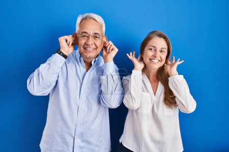 Photo for Middle age hispanic couple standing over blue background smiling pulling ears with fingers, funny gesture. audition problem - Royalty Free Image