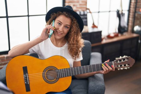 Photo for Young beautiful hispanic woman musician playing classical guitar talking on smartphone at music studio - Royalty Free Image