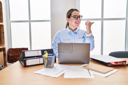 Foto de Joven mujer hispana trabajando en la oficina con gafas sonriendo con la cara feliz mirando y señalando hacia un lado con el pulgar hacia arriba. - Imagen libre de derechos