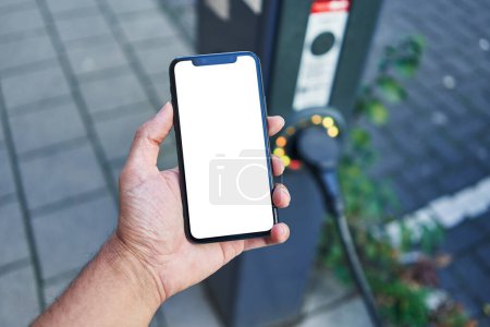 Foto de Man holding smartphone showing white blank screen at electrical car station - Imagen libre de derechos