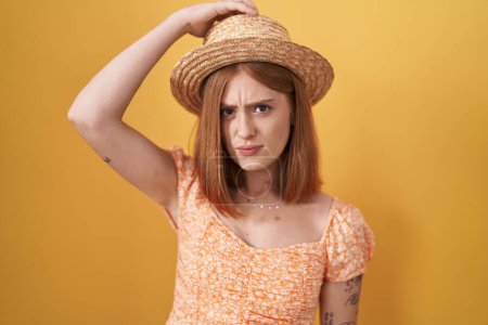 Photo for Young redhead woman standing over yellow background wearing summer hat confuse and wonder about question. uncertain with doubt, thinking with hand on head. pensive concept. - Royalty Free Image