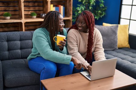 Foto de African american women friends drinking coffee using laptop at home - Imagen libre de derechos