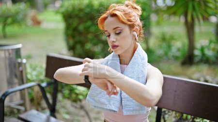 Téléchargez les photos : Jeune rousse femme utilisant smartwatch et écouteurs assis sur le banc au parc - en image libre de droit