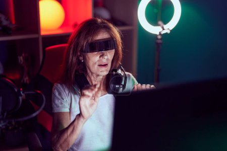 Téléchargez les photos : Femme du moyen âge streamer jouer à un jeu vidéo en utilisant des lunettes de réalité virtuelle à la salle de jeux - en image libre de droit