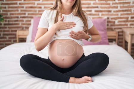 Téléchargez les photos : Jeune femme enceinte assise sur le lit examinant le sein avec les mains à la chambre - en image libre de droit