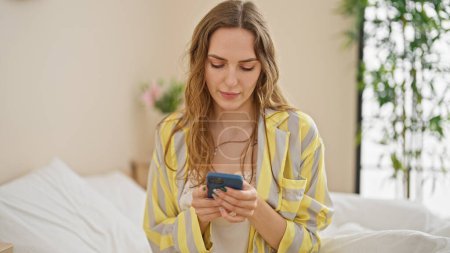 Foto de Mujer rubia joven usando teléfono inteligente sentado en la cama en el dormitorio - Imagen libre de derechos