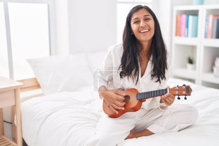 Photo for Young beautiful hispanic woman playing ukulele sitting on bed at bedroom - Royalty Free Image