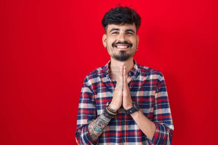 Photo for Young hispanic man with beard standing over red background praying with hands together asking for forgiveness smiling confident. - Royalty Free Image