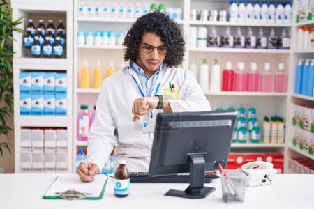 Téléchargez les photos : Homme hispanique avec les cheveux bouclés travaillant à la pharmacie pharmacie en regardant le temps de la montre inquiet, peur de se faire tard - en image libre de droit