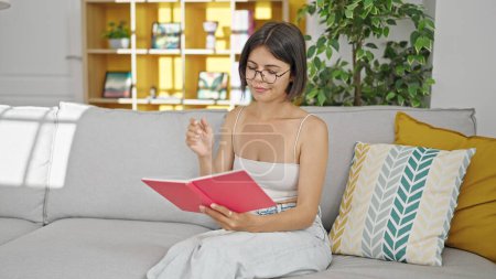 Photo for Young beautiful hispanic woman reading book sitting on sofa at home - Royalty Free Image
